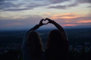 Two women, at sunset, holding their arms up in the air to create a small heart with their hands between them