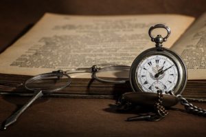 An old-fashioned pocket watch next to an open book and a pair of glasses